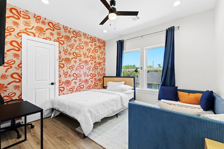 Chumba cha kulala kina ukuta wa rangi ya chungwa, kitanda chenye rangi ya chungwa, kitanda chenye starehe, kitanda cha mchana cha bluu, dawati la kazi, feni ya dari na madirisha makubwa yaliyo na mapazia ya baharini.