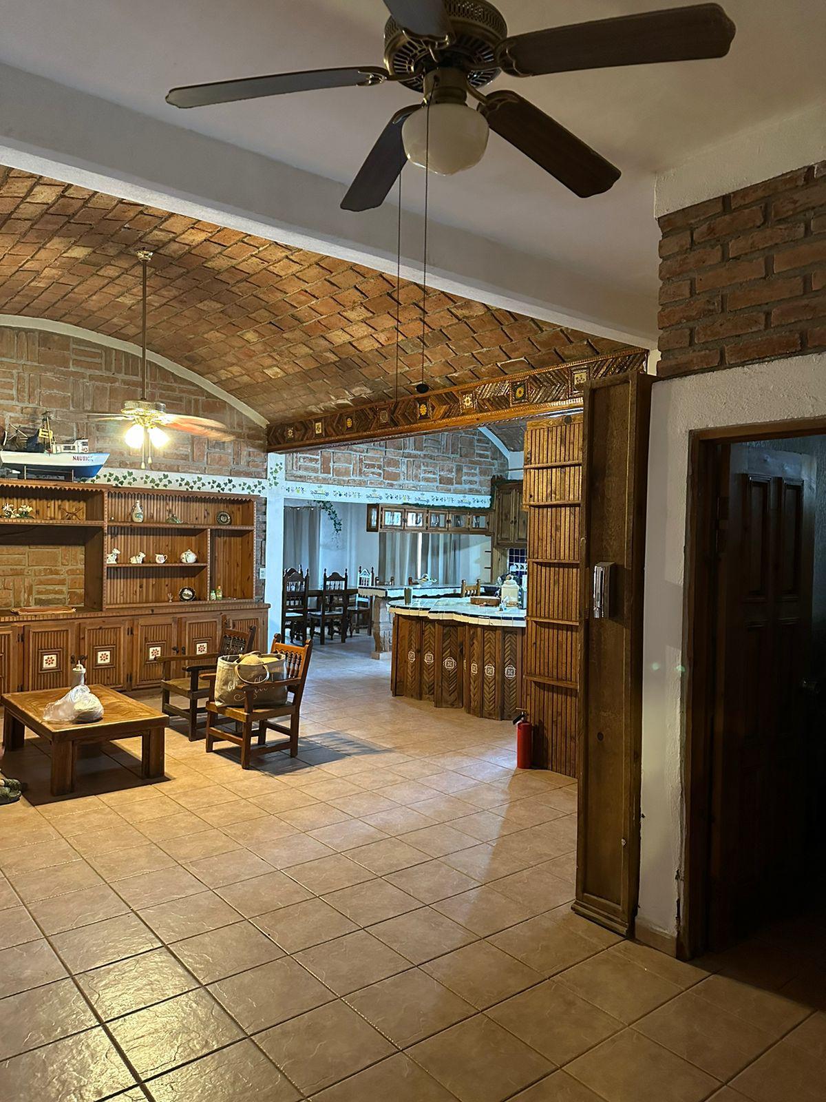 A spacious living area features tiled flooring and a high, arched ceiling. Wooden furniture, including a low table and several chairs, complements the warm brick walls. The adjoining kitchen is visible, showcasing a rustic design with wooden cabinetry and an open layout, enhancing communal living.