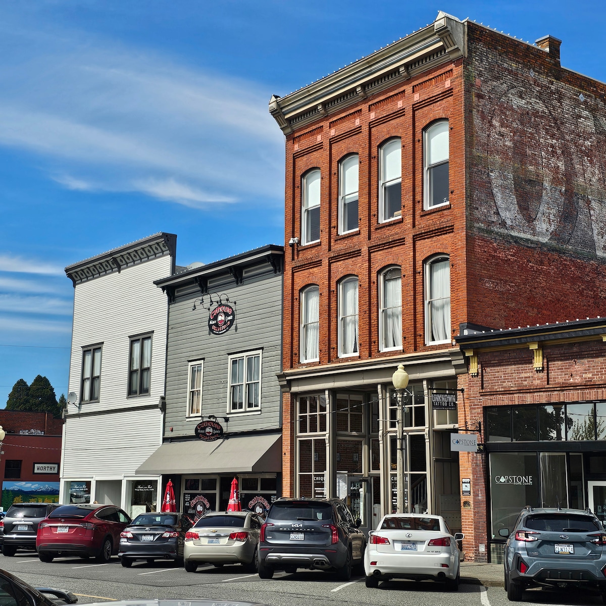 A bustling street scene is depicted, featuring a variety of businesses housed in historic buildings. The red brick structure stands prominently with large windows, while adjacent gray buildings feature outdoor seating. Several parked cars line the street, complementing the vibrant atmosphere.