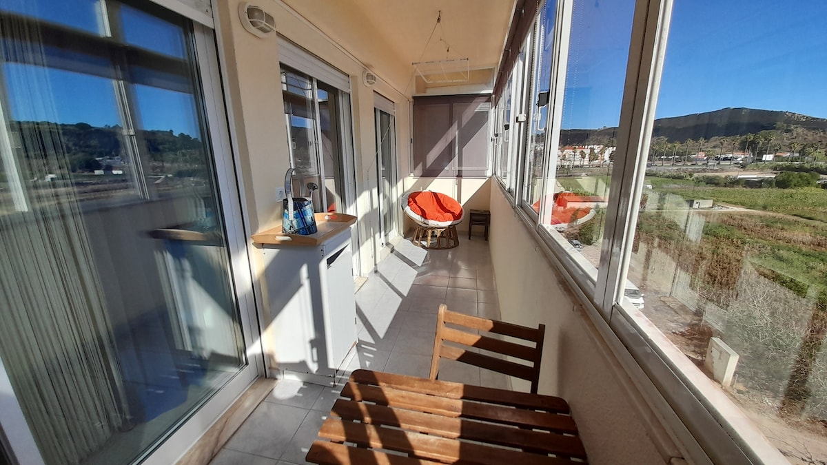 A spacious sunlit balcony features large windows providing panoramic views of the surrounding landscape. A small wooden table and two chairs are positioned on the tiled floor, alongside a cozy orange chair. The area is bathed in natural light, enhancing the inviting atmosphere.