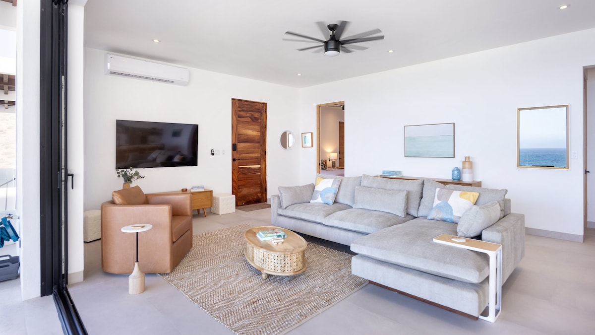 A spacious living area features a large sectional sofa arranged around a round coffee table, with light streaming in through floor-to-ceiling sliding glass doors. A wall-mounted television and a cozy accent chair enhance the inviting atmosphere, while a wood door leads to additional rooms.