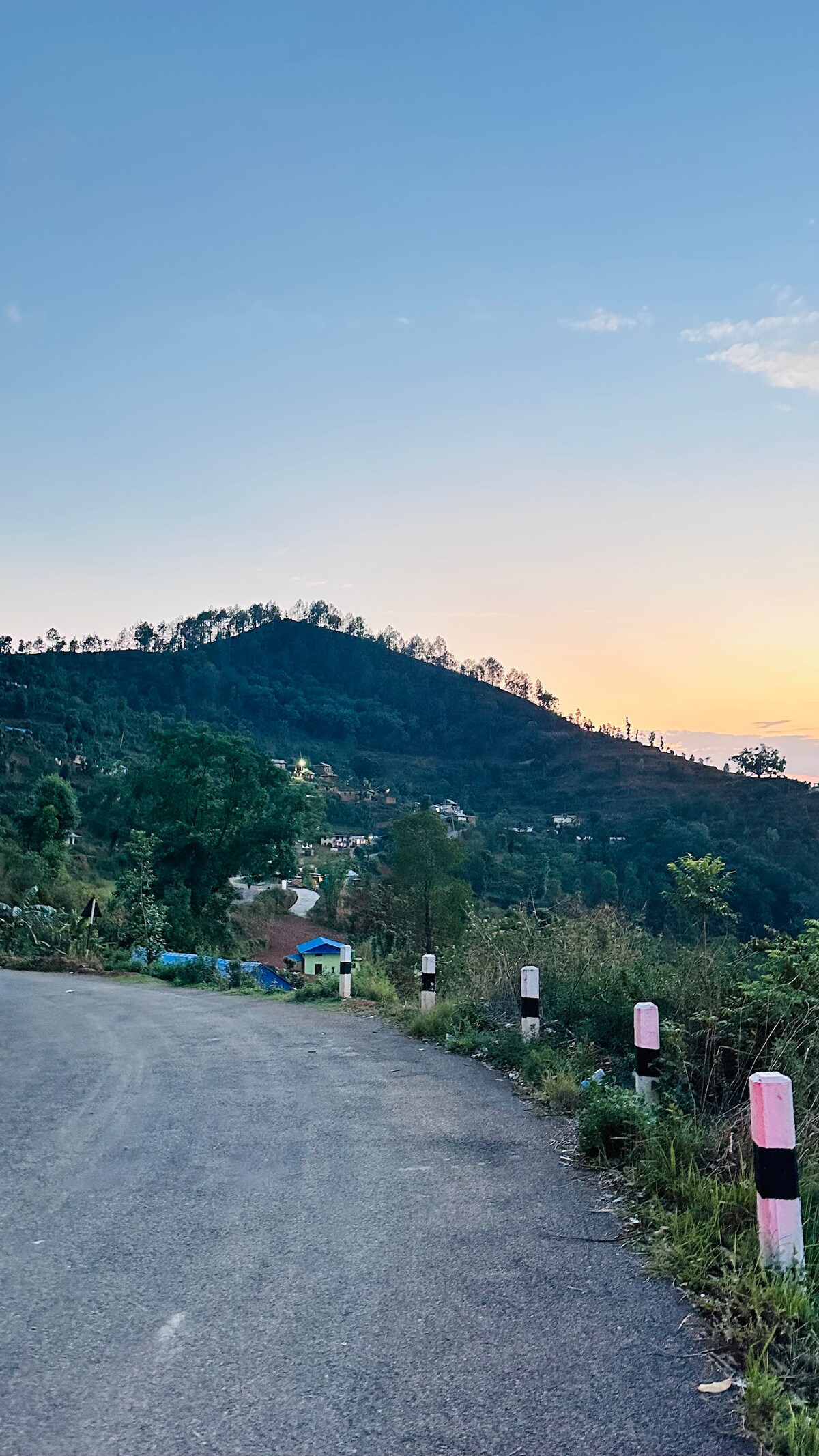 A scenic view of a winding road leads towards a hillside adorned with lush greenery. The sun sets in the background, casting warm hues into the sky, while colorful houses can be seen nestled within the valley, contributing to the tranquil landscape.