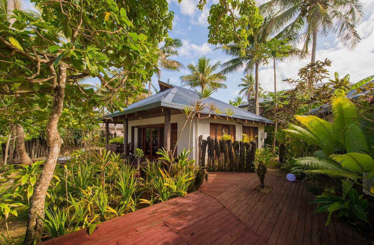 The villa exterior is surrounded by lush tropical greenery, with vibrant palm trees and plants framing the entrance. A wooden pathway leads up to the entrance, complemented by a covered porch with multiple windows and natural light filtering through.