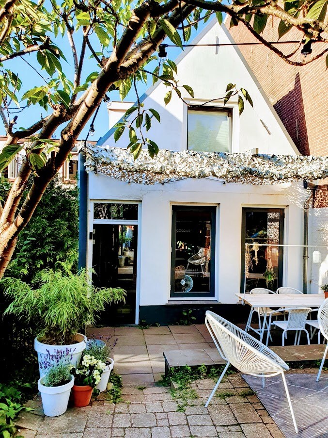 A charming patio area is enclosed by greenery, featuring a dining set for leisurely meals. The space includes a stone pathway and flower pots with various plants. Sunlight filters through a canopy, illuminating the white exterior of the home and large windows.