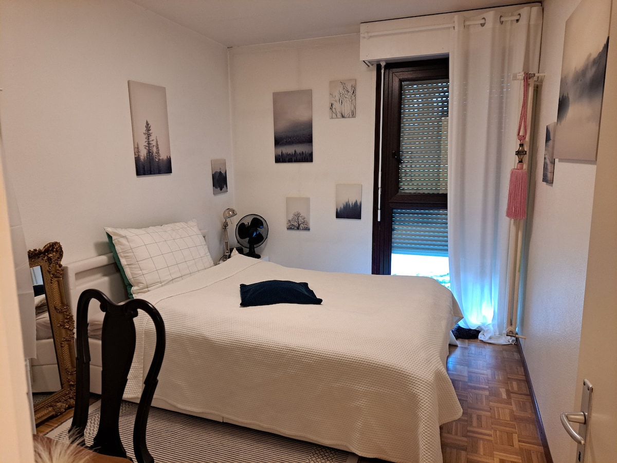 A bright bedroom features a spacious bed with a white bedspread and a decorative pillow. The room is adorned with wall art depicting serene landscapes. A window with sheer curtains allows natural light, while a small fan is positioned nearby. A framed mirror and a dark chair add to the decor.