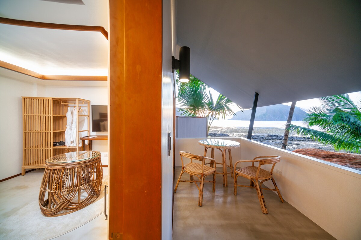 A private balcony is showcased with a round glass table and two woven chairs, surrounded by palm trees. In the background, the ocean can be seen, enhancing the serene setting. The adjacent indoor area includes a TV and a bamboo wardrobe, reflecting modern design.