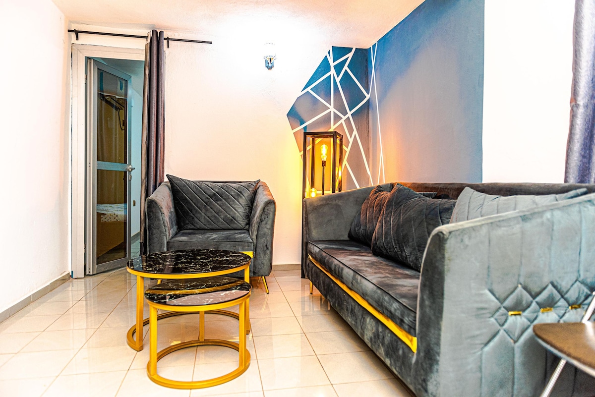 A stylish living area features a dark gray velvet sofa and a matching armchair, positioned around a circular black and gold coffee table. A geometric accent wall in blue adds visual interest, while soft lighting creates a warm ambiance. Dark curtains frame a nearby entrance.