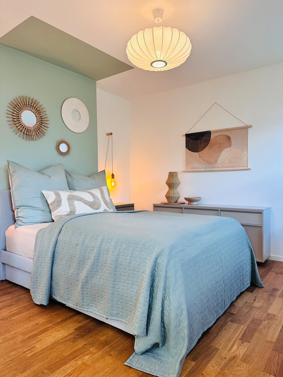 A cozy sleeping area features a spacious bed dressed in a light blue quilt. The walls are adorned with decorative round mirrors and a piece of wall art. Soft lighting is provided by a pendant lamp, highlighting a warm wooden floor and minimalist furniture.