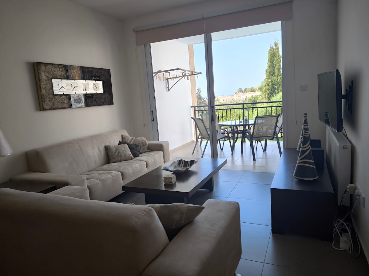 A comfortable living area is highlighted by a light-colored sofa and an accent chair, arranged around a low table. A wall-mounted television is positioned across from the seating. Large glass doors provide access to a balcony with outdoor seating, overlooking greenery.