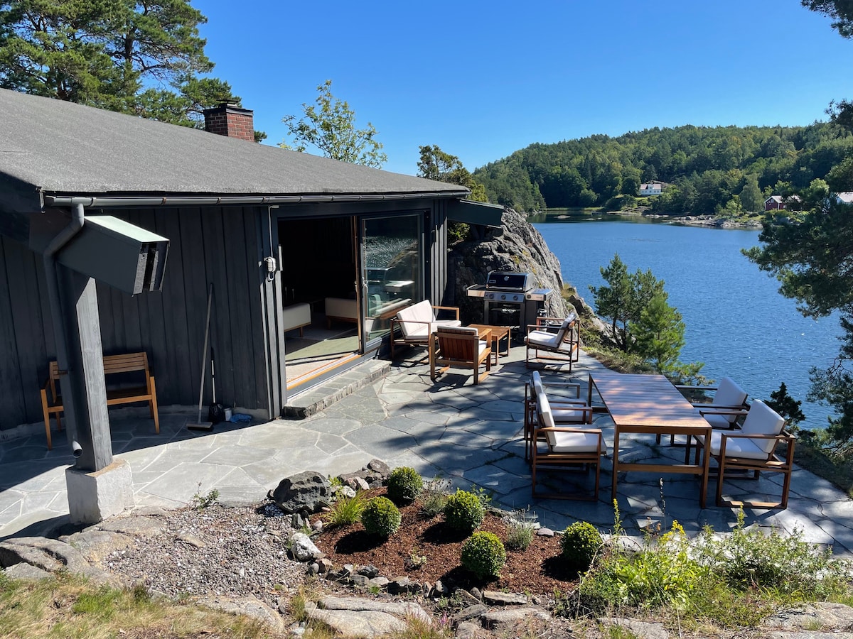 An outdoor patio is featured with multiple wooden seating arrangements, surrounded by landscaped greenery and rocky terrain. A view of the water is visible beyond the house, while a barbecue grill is positioned nearby. Sunlight illuminates the area, creating an inviting space for relaxation.