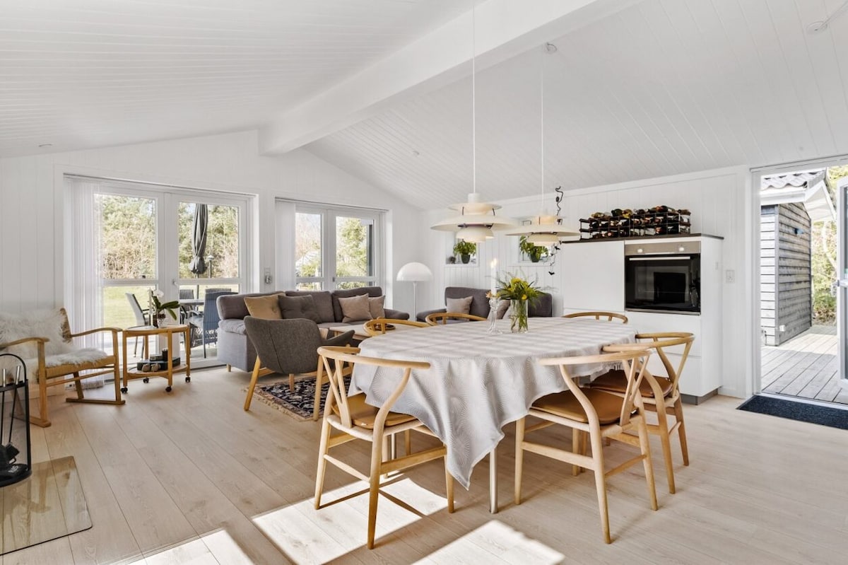 An open-plan living and dining area features a large wooden table surrounded by light chairs. Cozy seating arrangements with gray sofas are present, and large windows allow natural light to fill the space. A subtle decor of indoor plants and a rug enhances the ambiance.