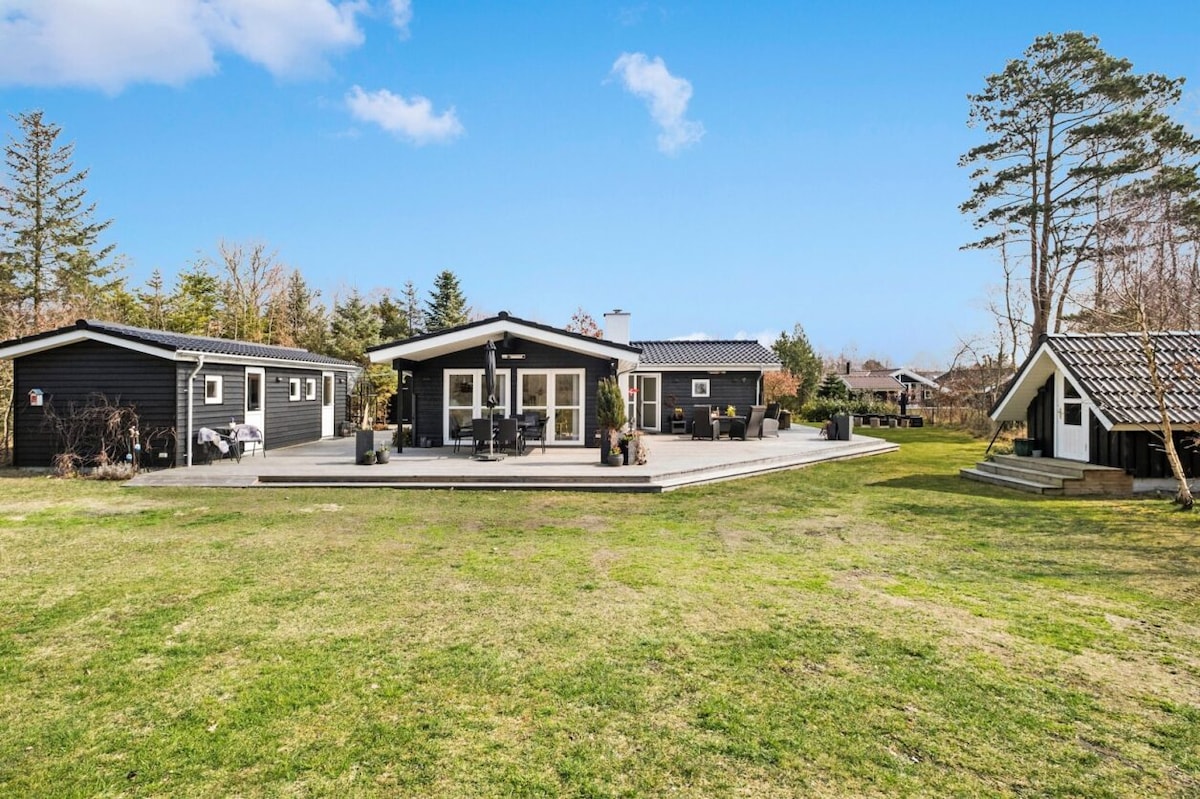 A spacious house is surrounded by a well-maintained lawn, featuring a large wooden deck with seating arrangements. Adjacent structures, including an outbuilding, are visible, set against a backdrop of trees and open sky.