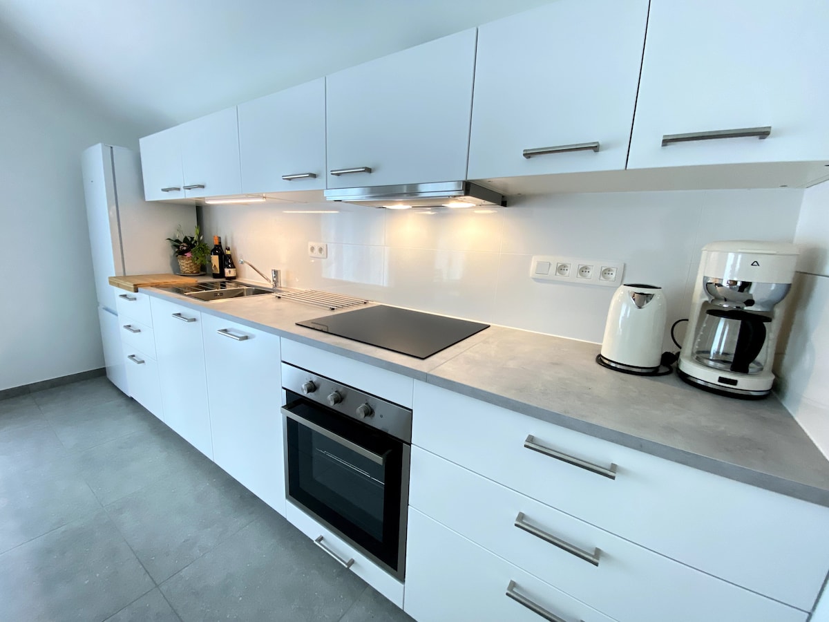 A modern kitchen is displayed, featuring sleek white cabinetry and a functional layout. Counter space includes a stove, oven, and coffee maker. Natural light brightens the gray tile flooring, and plants are placed on the counter for a touch of greenery.