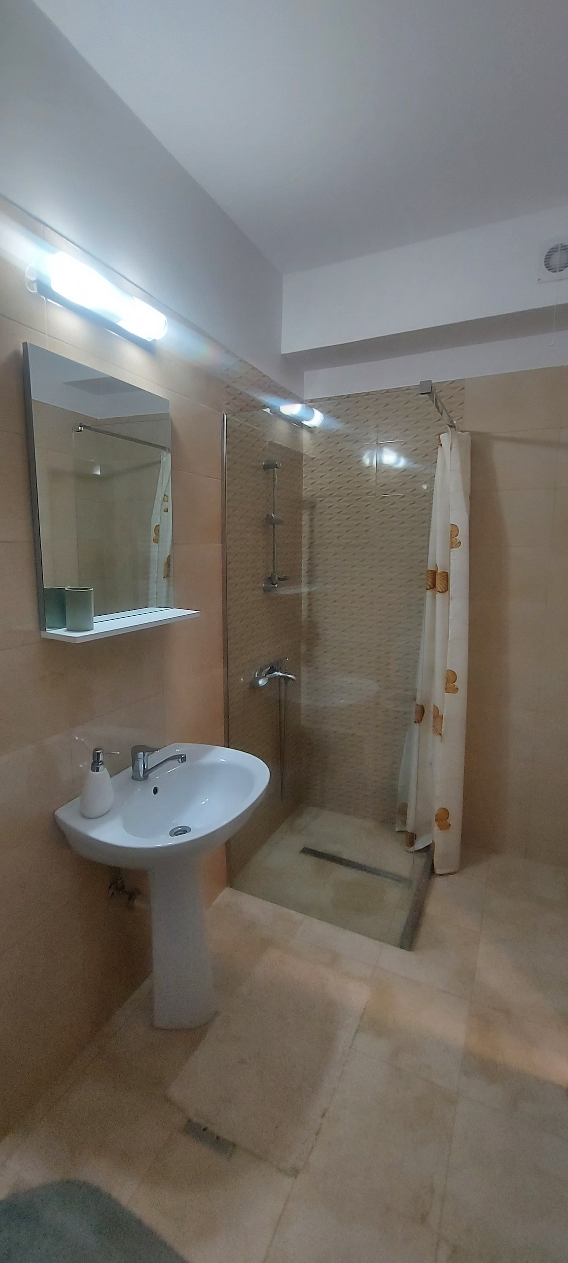 A modern bathroom features a walk-in shower enclosed by transparent glass, complemented by a stylish showerhead. A round mirror above a pedestal sink reflects ample lighting from overhead fixtures. The neutral tile colors create a clean and spacious environment, enhancing the functionality of the area.