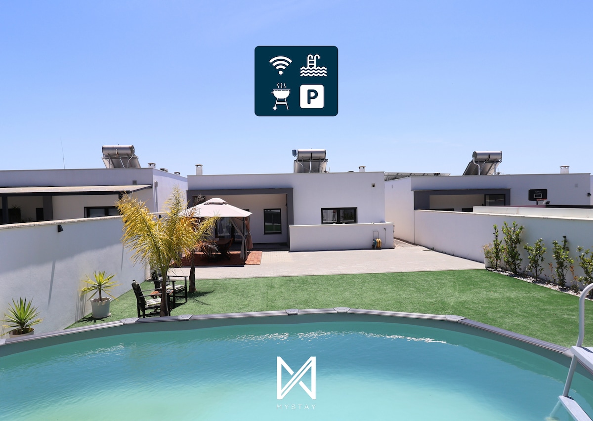 A clear view of a private pool surrounded by a well-maintained garden. The outdoor dining area, equipped with a barbecue, is situated on a spacious patio. The house features modern architecture, with a light-colored exterior and large windows allowing natural light.