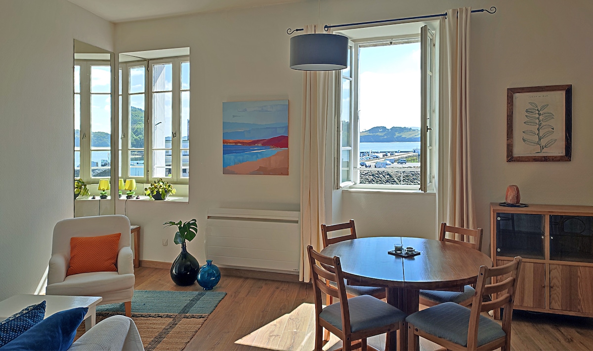 A bright living area features large windows that offer views of the harbor. A round wooden table is surrounded by chairs, while a cozy armchair is positioned nearby. Soft colors adorn the walls, complemented by artwork and decorative plants on the window sill.