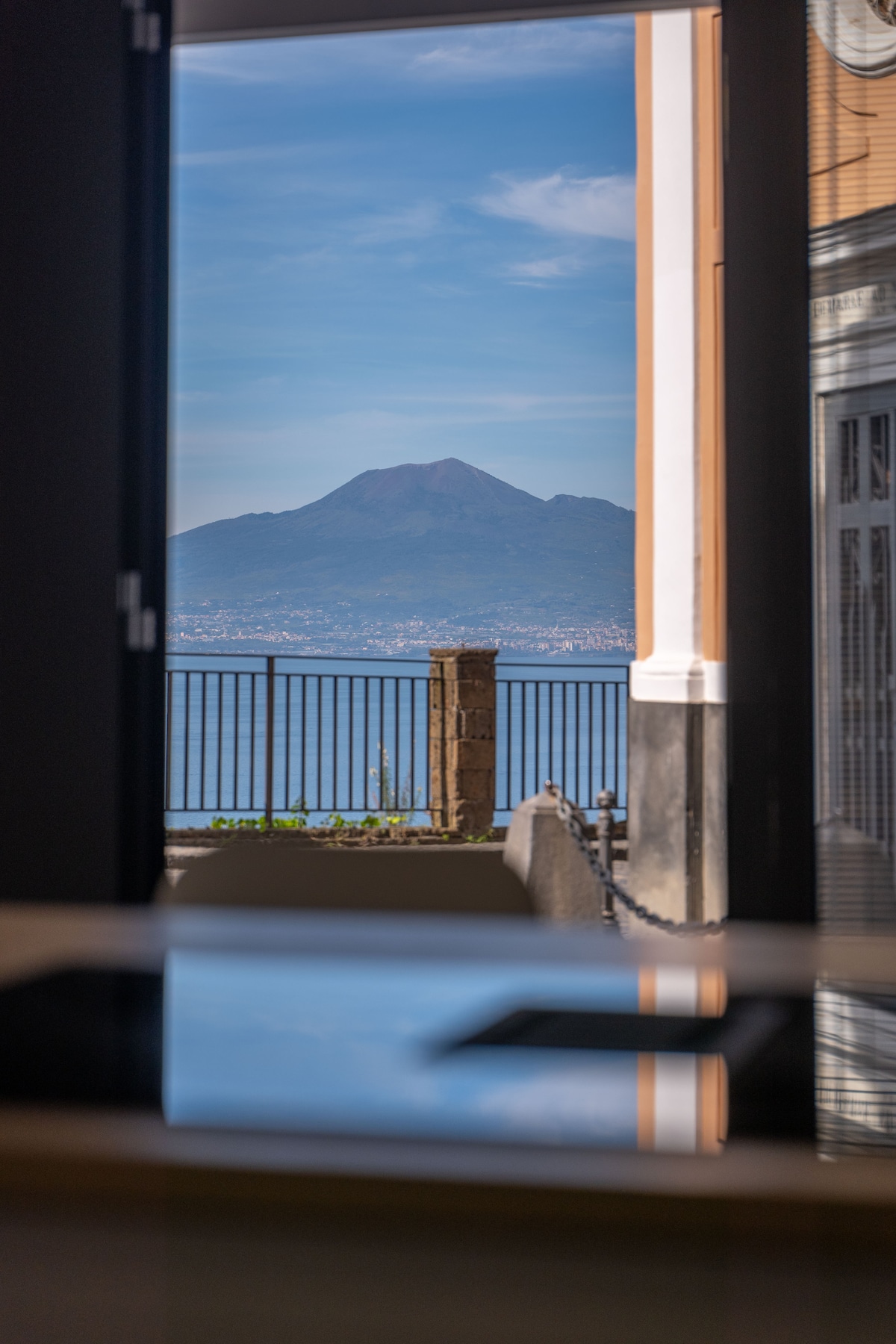 The image showcases a view of Mount Vesuvius through an open doorway. A wrought iron railing is visible in the foreground, framing the landscape with the mountain against a clear blue sky. Reflections on a surface hint at the interior space.