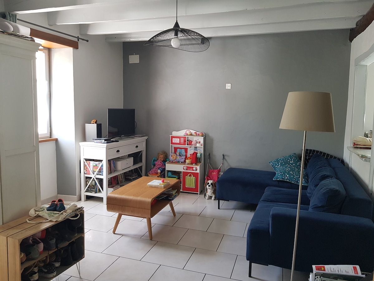 A living area features a comfortable blue sectional sofa and a stylish wooden coffee table. A television is positioned on a white media console, surrounded by shelves holding various decorative items. Light-colored tiles cover the floor, and a lamp stands beside the sofa, adding illumination.
