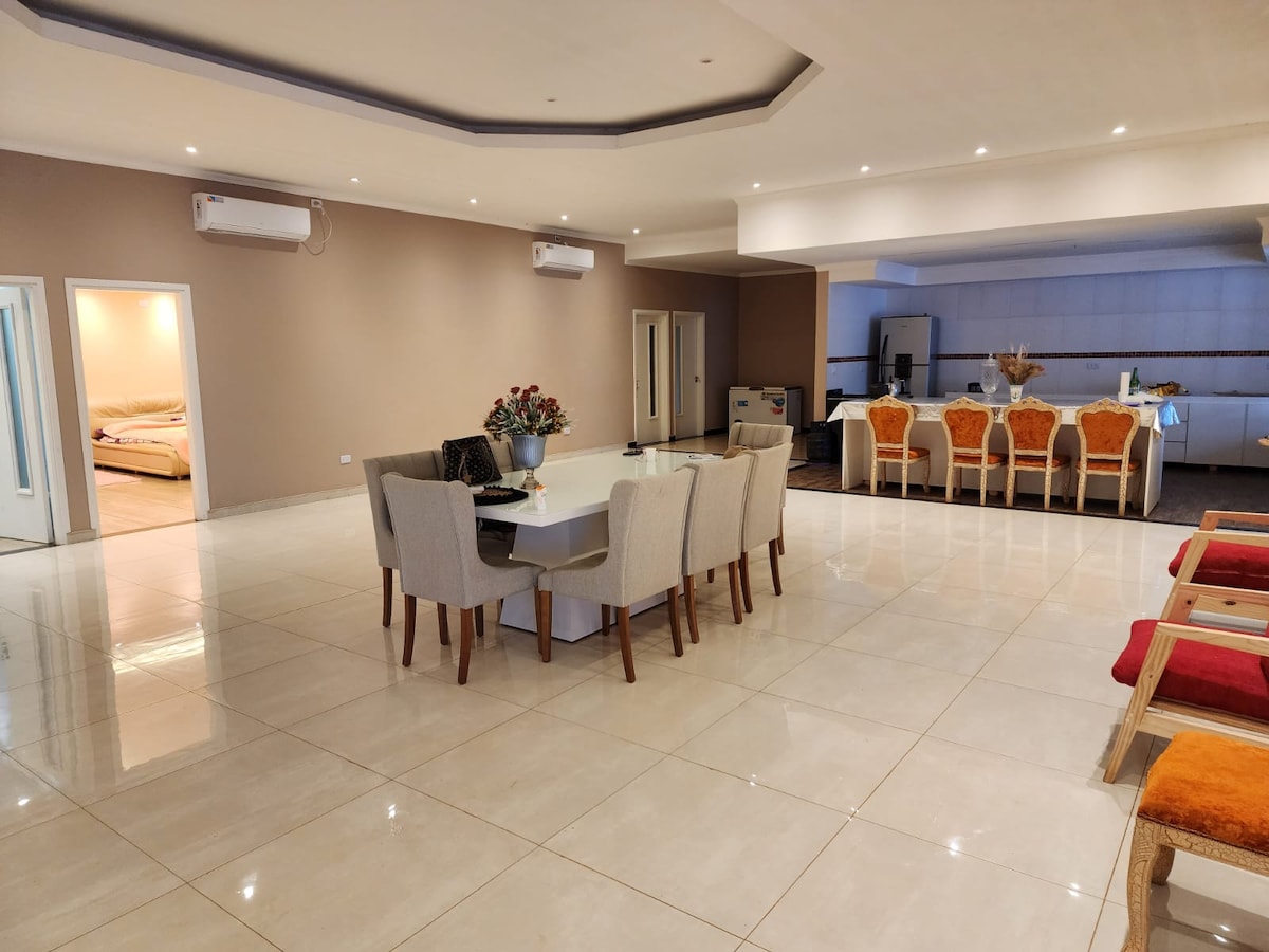 A spacious dining area features a large table surrounded by cream-colored chairs. Brightly lit by overhead lighting, the room showcases sleek white tiles. In the background, an open kitchen is visible, with a counter and several orange upholstered chairs, enhancing the versatile layout.