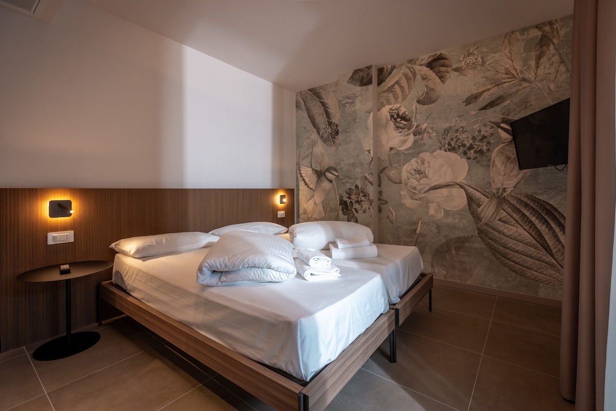 A modern bedroom features a spacious bed with plush pillows and soft linens. Decorative floral wallpaper adds a touch of elegance to the room. A minimalist nightstand is positioned beside the bed, and a wall-mounted TV is visible in the corner.