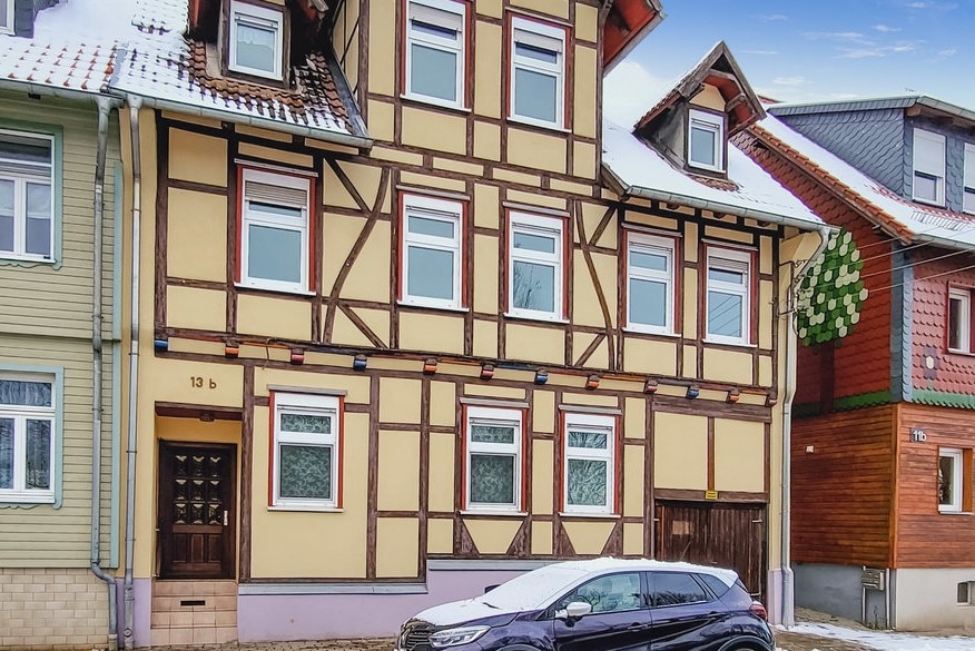 A historic timber-framed building is presented, featuring an array of windows adorned with white trim. The exterior retains a classic charm, with a blend of yellow and brown hues. A parked vehicle is visible in front of the house, while snow covers the ground.