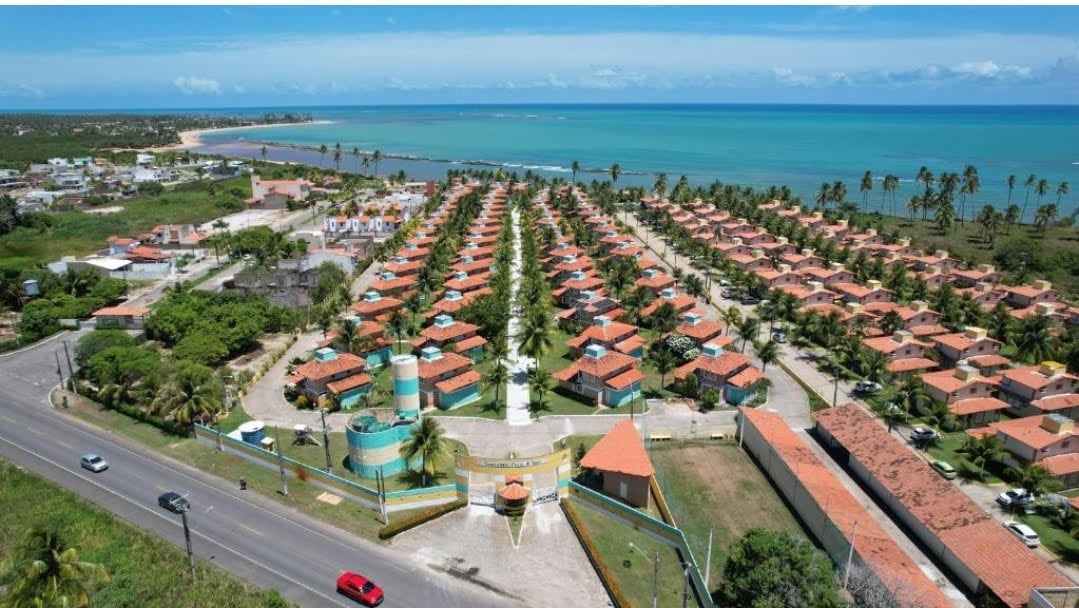 An aerial view captures a residential complex with several single-story houses featuring red roofs. A well-maintained road borders the property, leading towards a scenic coastline with turquoise waters and palm trees swaying along the beach.