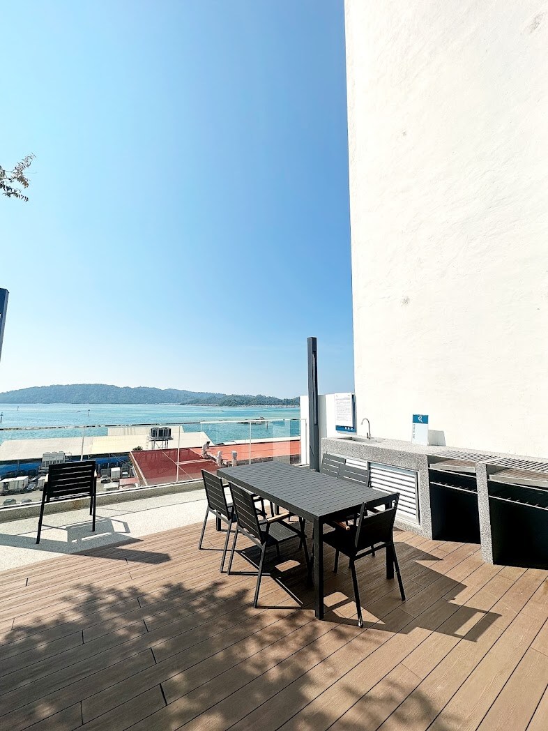 An outdoor terrace is shown featuring a black dining table with matching chairs. The area is surrounded by a railing, showcasing a scenic view of the sea and distant land, illuminated by bright sunlight. A small sink is visible, enhancing the outdoor functionality.