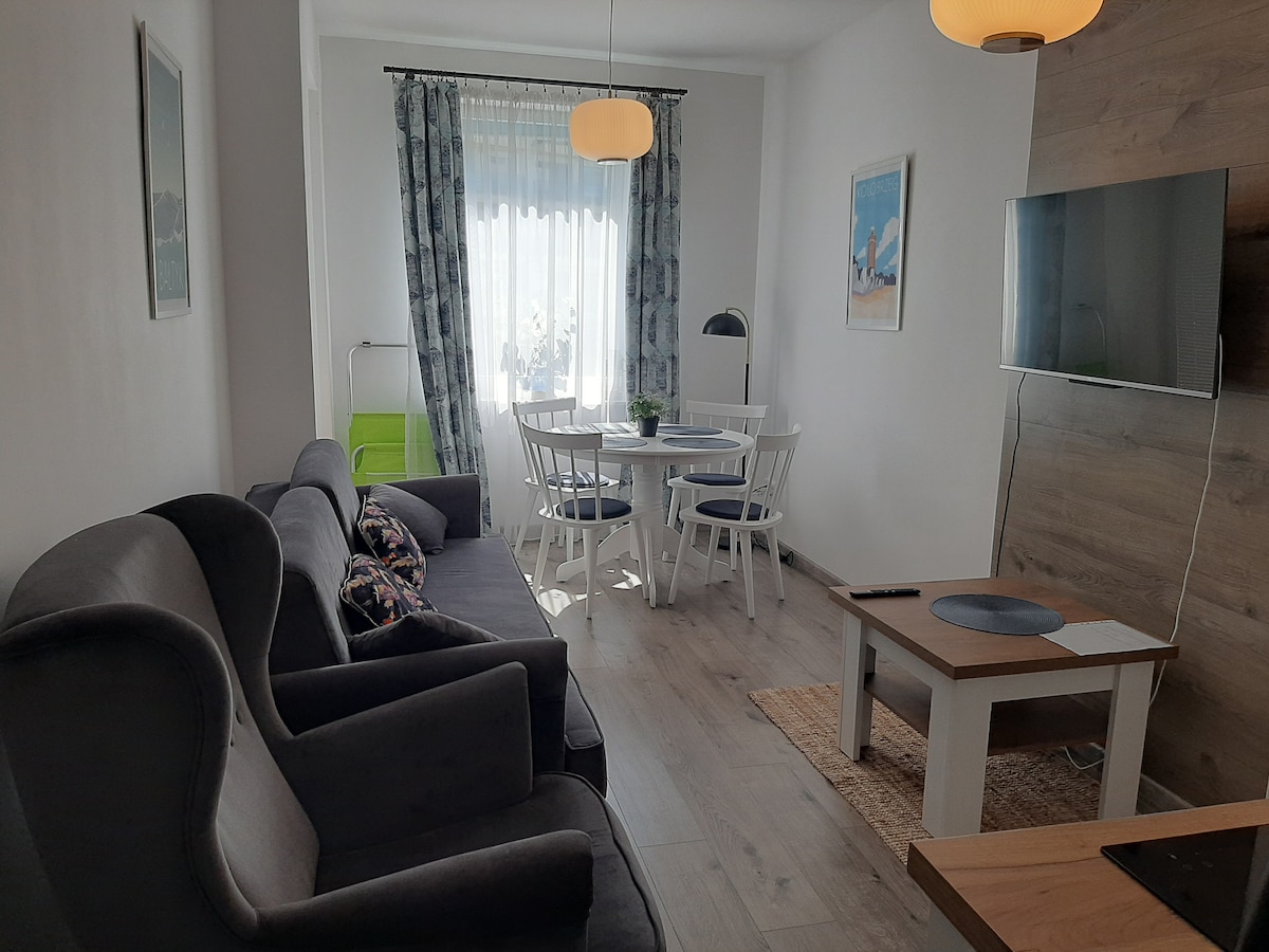 A bright living area is presented, featuring a neutral color palette. Two comfortable armchairs are positioned around a low coffee table. A round dining table with four chairs is illuminated by natural light through a window with sheer curtains. A television is mounted on the wall.