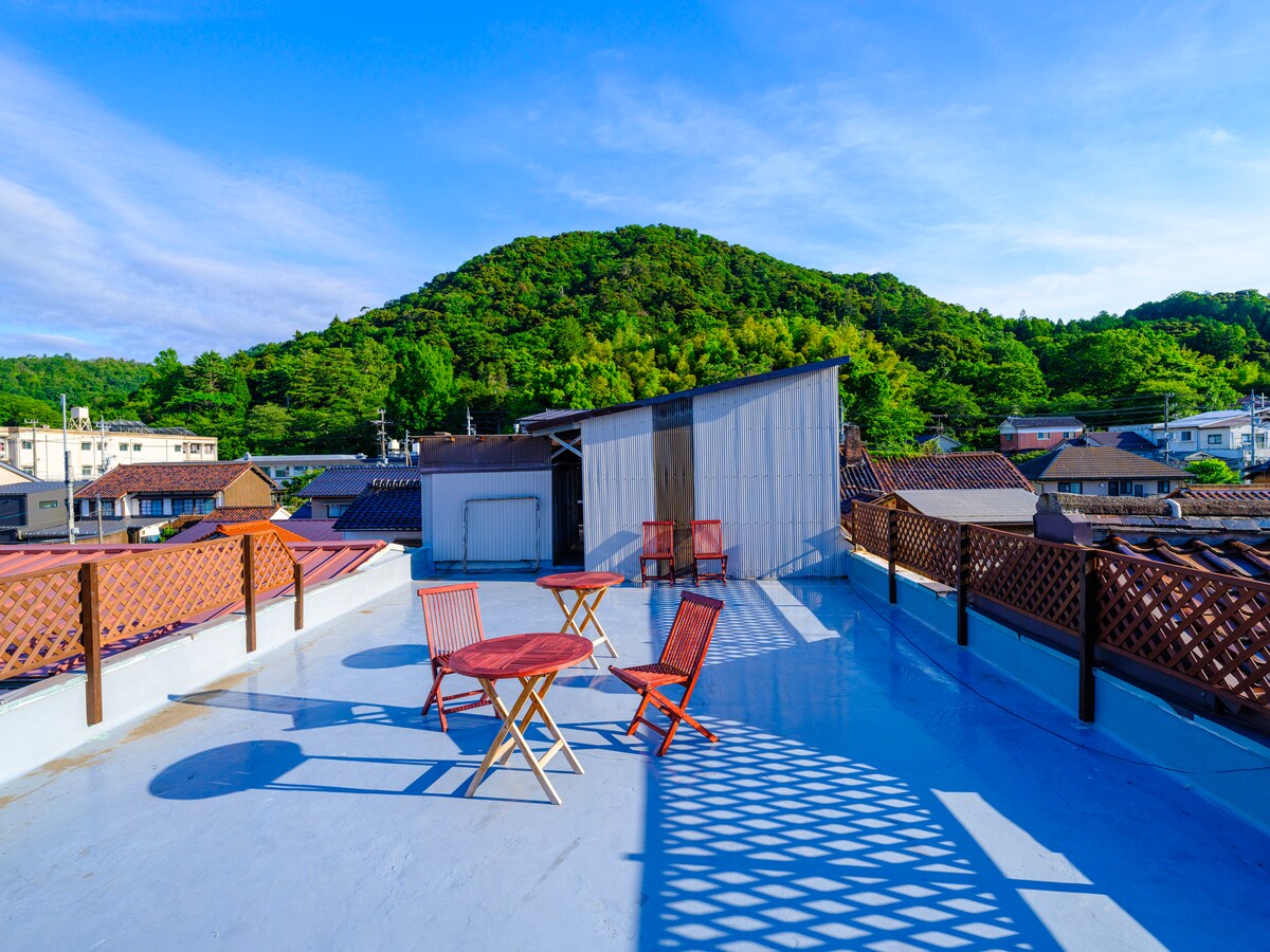 A rooftop terrace is showcased, featuring a smooth blue surface and a panoramic view of the surrounding hillside. Two folding tables and four chairs are arranged, providing a space for relaxation amidst the scenic landscape.
