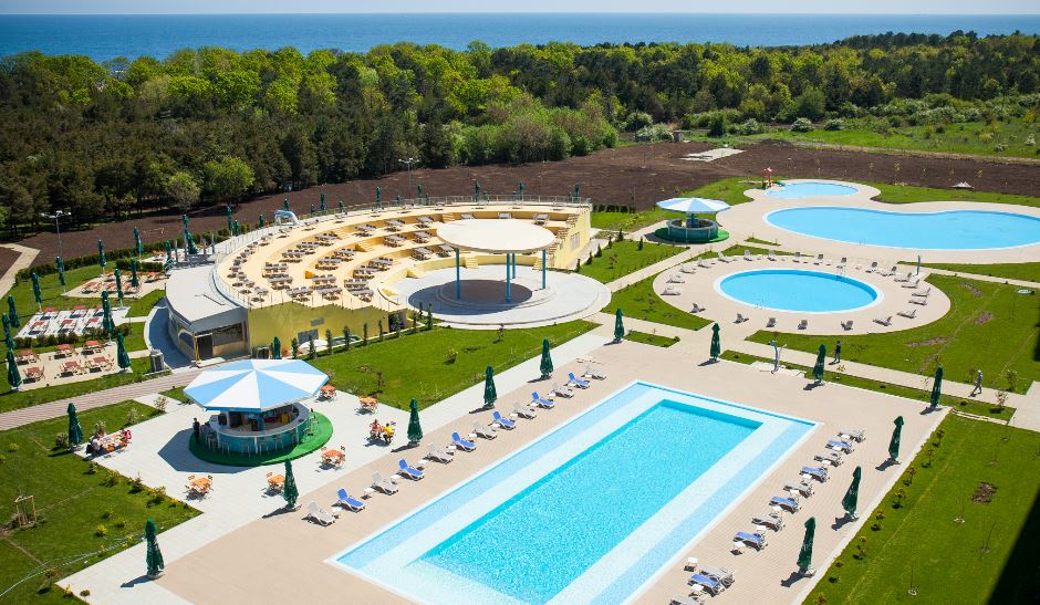 A spacious outdoor area is depicted, featuring three swimming pools surrounded by green lawns. Sun loungers are neatly arranged along the pool edges. A shaded bar area with seating is visible, along with a large circular structure that may serve as a communal space.