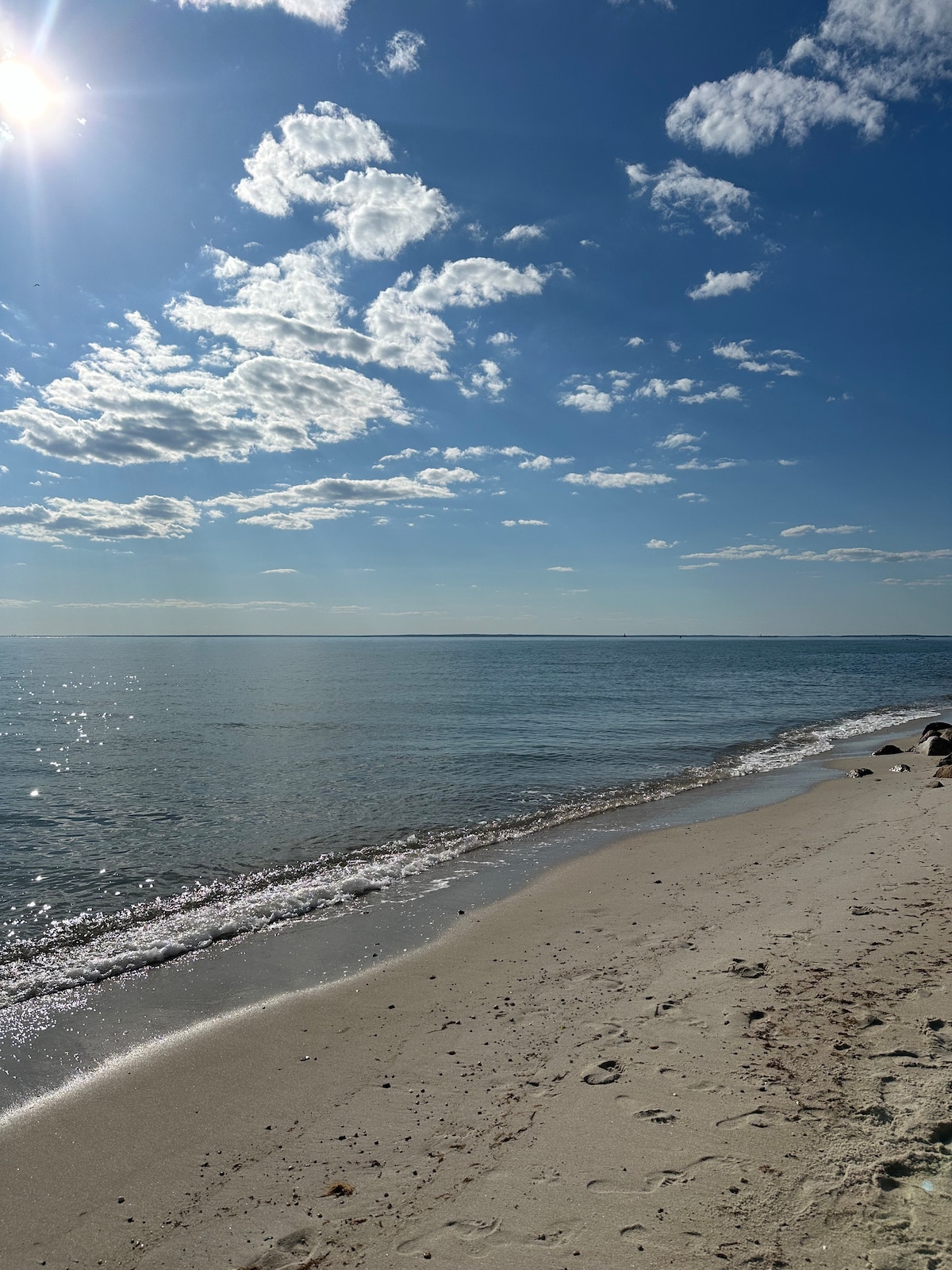 A sandy beach stretches along a calm shoreline, where gentle waves meet the sand. The clear blue sky is dotted with soft white clouds, and sunlight reflects off the water's surface, creating a serene atmosphere.
