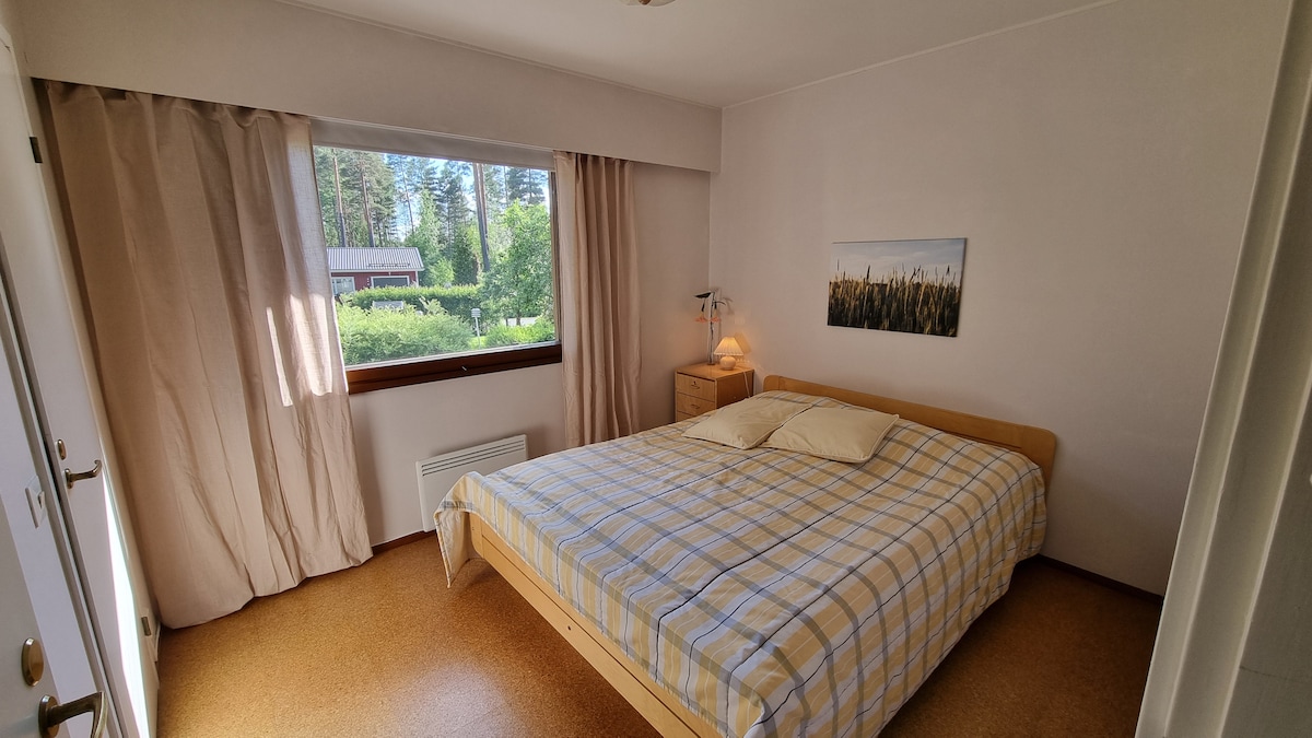 A bright guest room features a spacious bed dressed in a light checkered blanket. Soft curtains frame a large window, allowing natural light to illuminate the space. A bedside lamp and a small nightstand add functionality, while a serene view of greenery is visible outside.