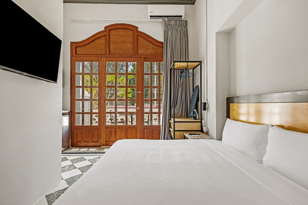 A spacious room features a large bed with white linens, a wall-mounted television, and a wardrobe. Expansive glass doors lead to a private outdoor area, while soft curtains frame the window, allowing natural light to fill the space.