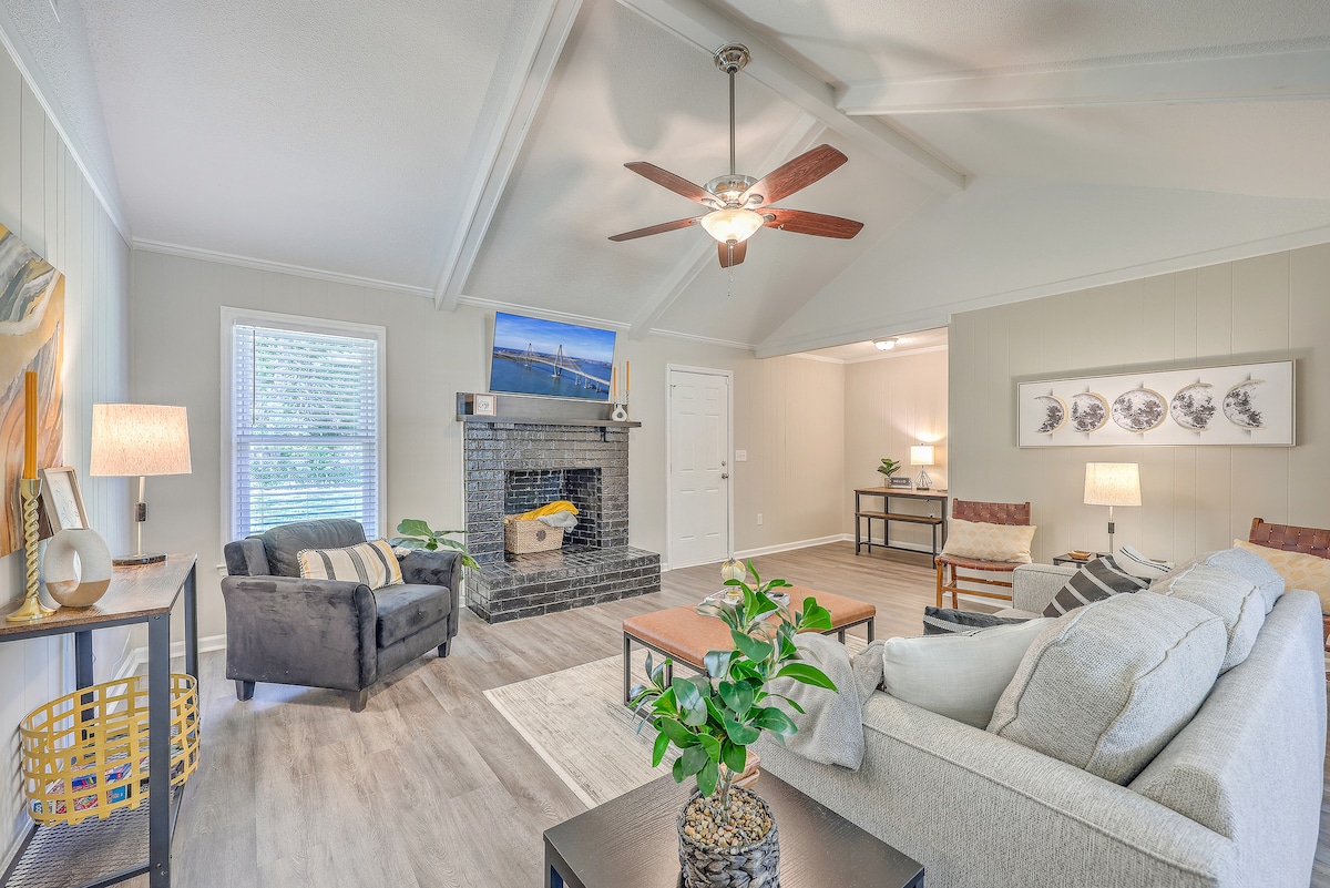 A bright living room features a cozy sofa and armchairs, arranged around a coffee table. Natural light enters through windows, illuminating the space. A decorative plant and lamp are visible on a side table. A ceiling fan and art piece above a fireplace enhance the inviting atmosphere.