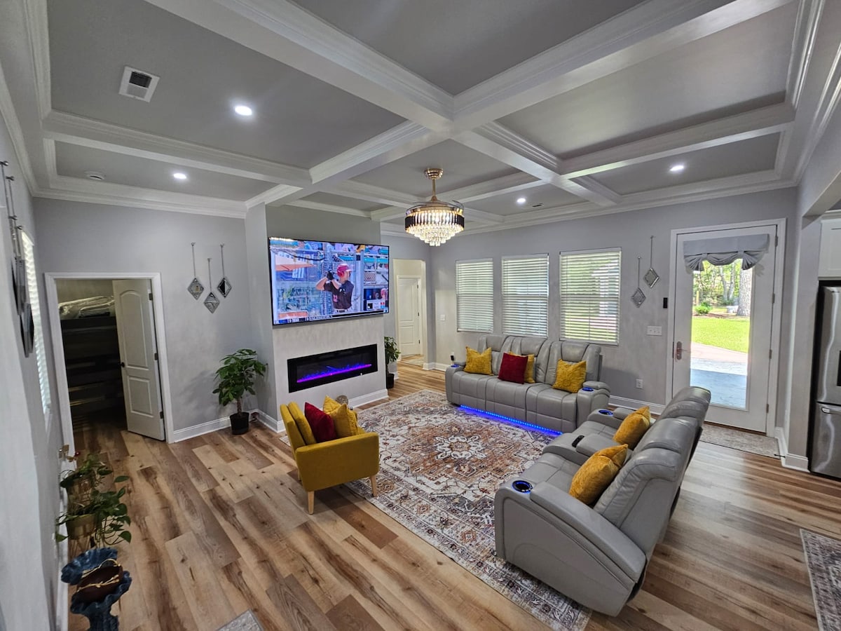 A spacious living area is highlighted by a modern sectional sofa with colorful cushions. A large television is mounted above an electric fireplace, providing a focal point within the room. Natural light streams in through multiple windows, illuminating the stylish decor and wooden flooring.