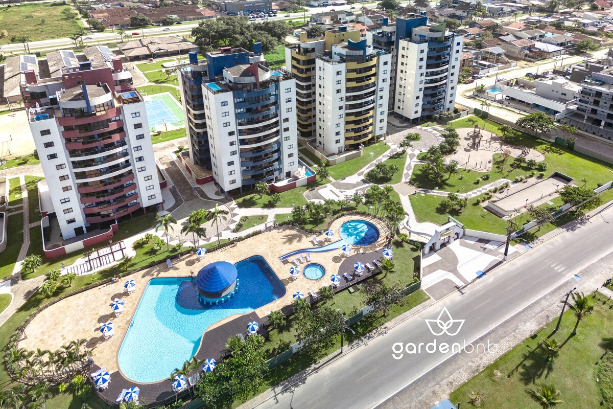 An aerial view of the condominium complex displays the expansive swimming pool surrounded by sun loungers. The lush green lawn provides ample outdoor space, while the nearby sports facilities and playground are visible, enhancing the community atmosphere.