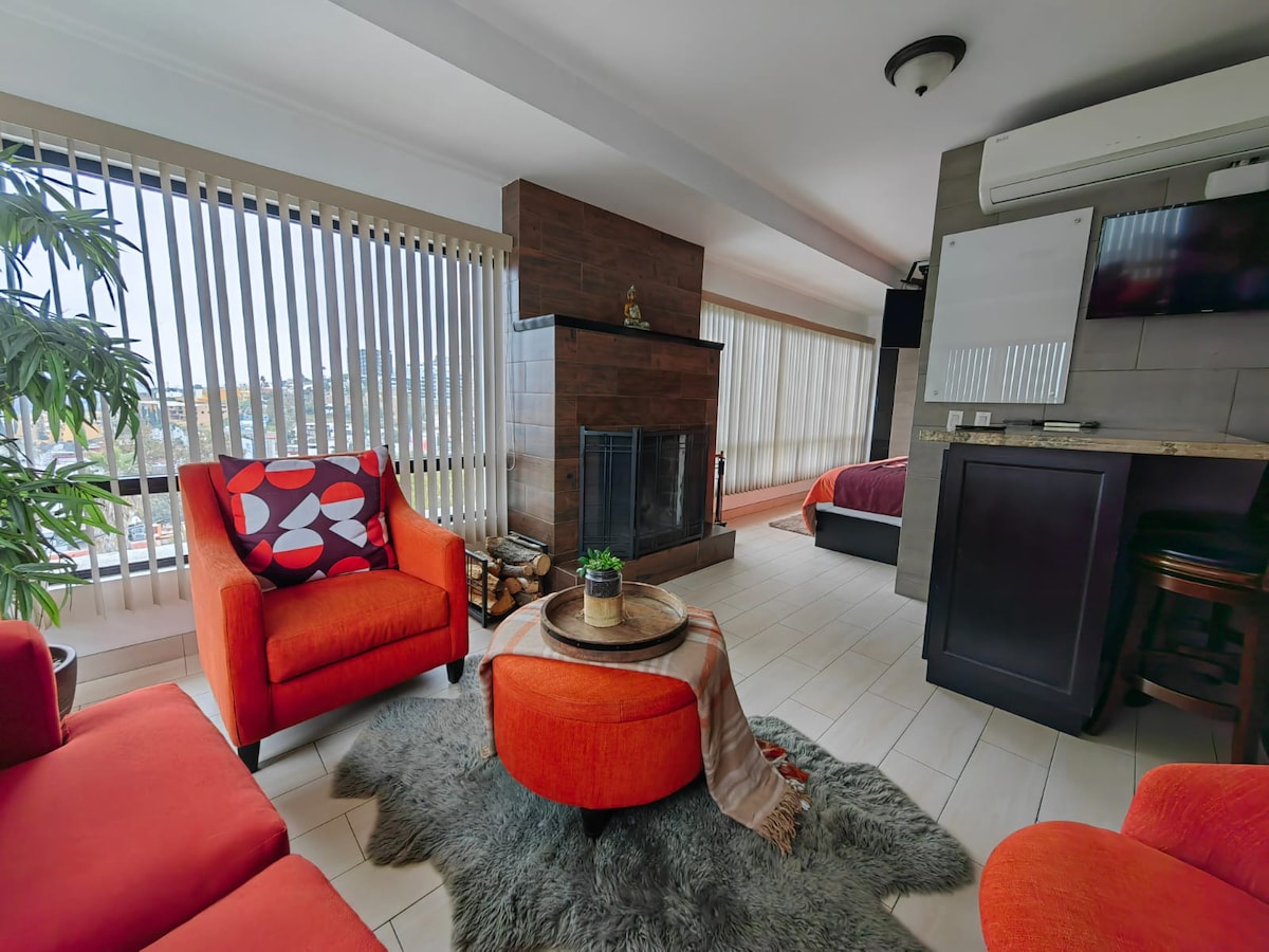 A modern living area features two vibrant red chairs positioned around a round coffee table. A cozy rug complements a stone fireplace, while large windows with vertical blinds allow natural light to brighten the space. In the background, the bedroom area is visible.