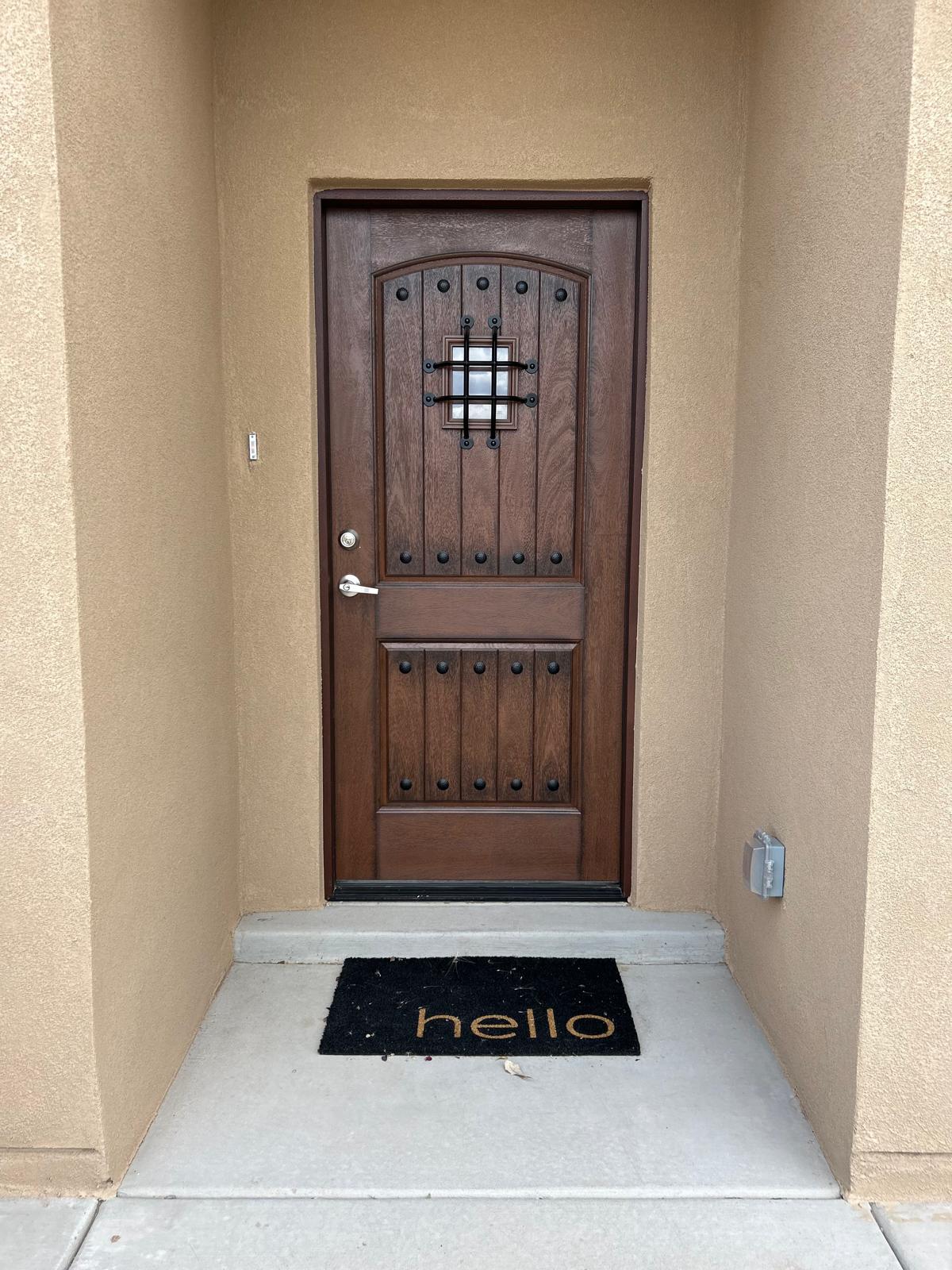 The entrance features a solid wooden door with decorative iron accents and a small window. A welcoming doormat with the word 'hello' is placed on the concrete entryway, which is surrounded by smooth stucco walls.