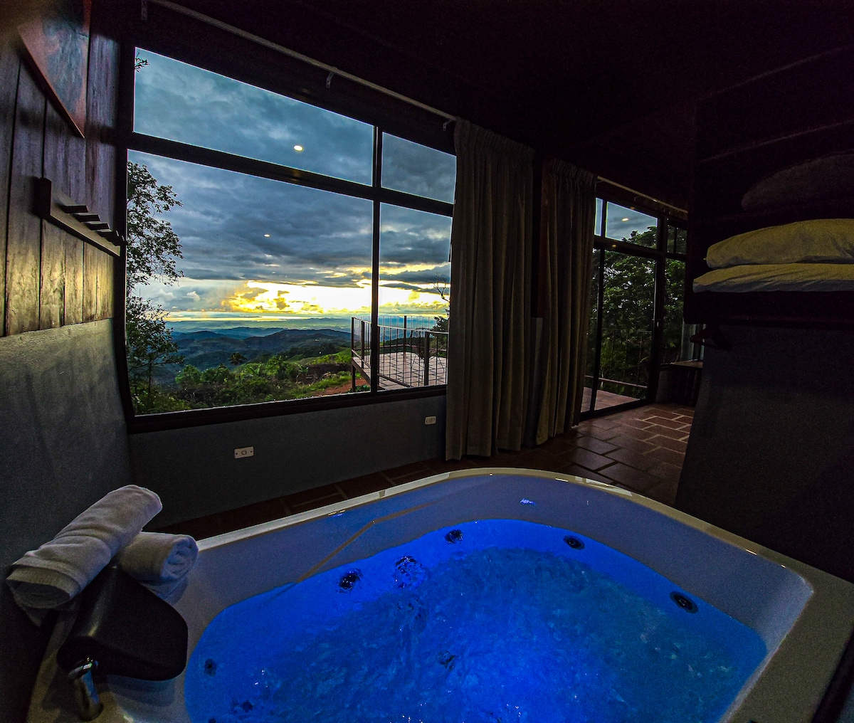 A relaxing hot tub is depicted in the foreground, illuminated with soft blue light. Large windows offer an expansive view of the landscape, featuring a cloudy sky and distant hills. Plush towels are neatly arranged beside the tub.