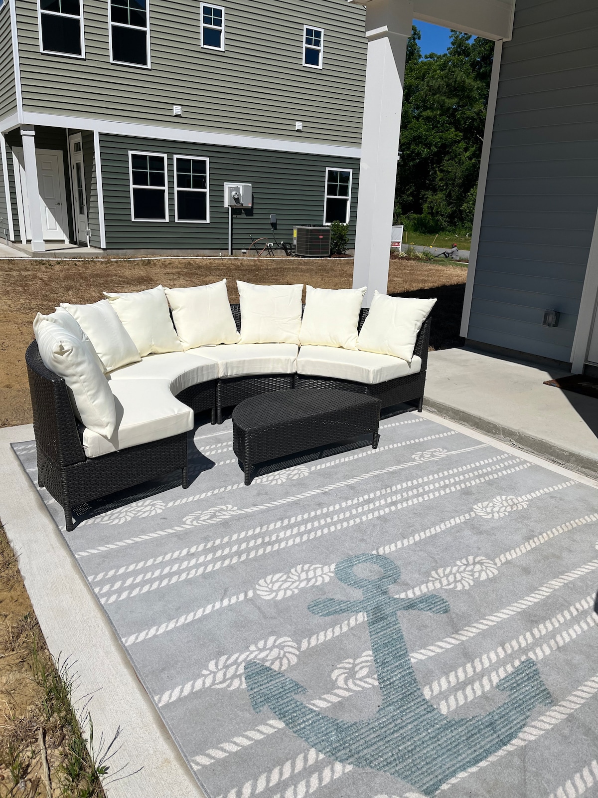 An outdoor seating area features a curved sectional sofa with light-colored cushions, arranged on a patterned area rug. The background displays a newly constructed home with several windows, and a gravel area can be seen adjacent to the patio.