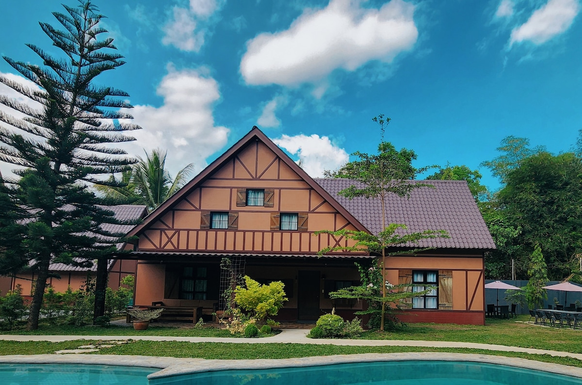 A spacious cabin is framed by lush greenery and blue skies. The triangular roof adds character, while large windows invite natural light. A clear swimming pool reflects the structure, and neatly arranged seating is visible on the surrounding lawn.