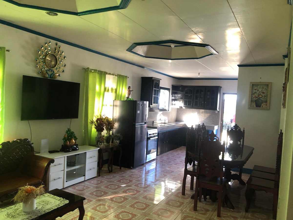 A welcoming living area features a large television mounted on the wall and a spacious dining table surrounded by wooden chairs. The kitchen area is visible, showcasing modern appliances. Bright green curtains frame the windows, allowing natural light to fill the space. Decorative elements add charm.