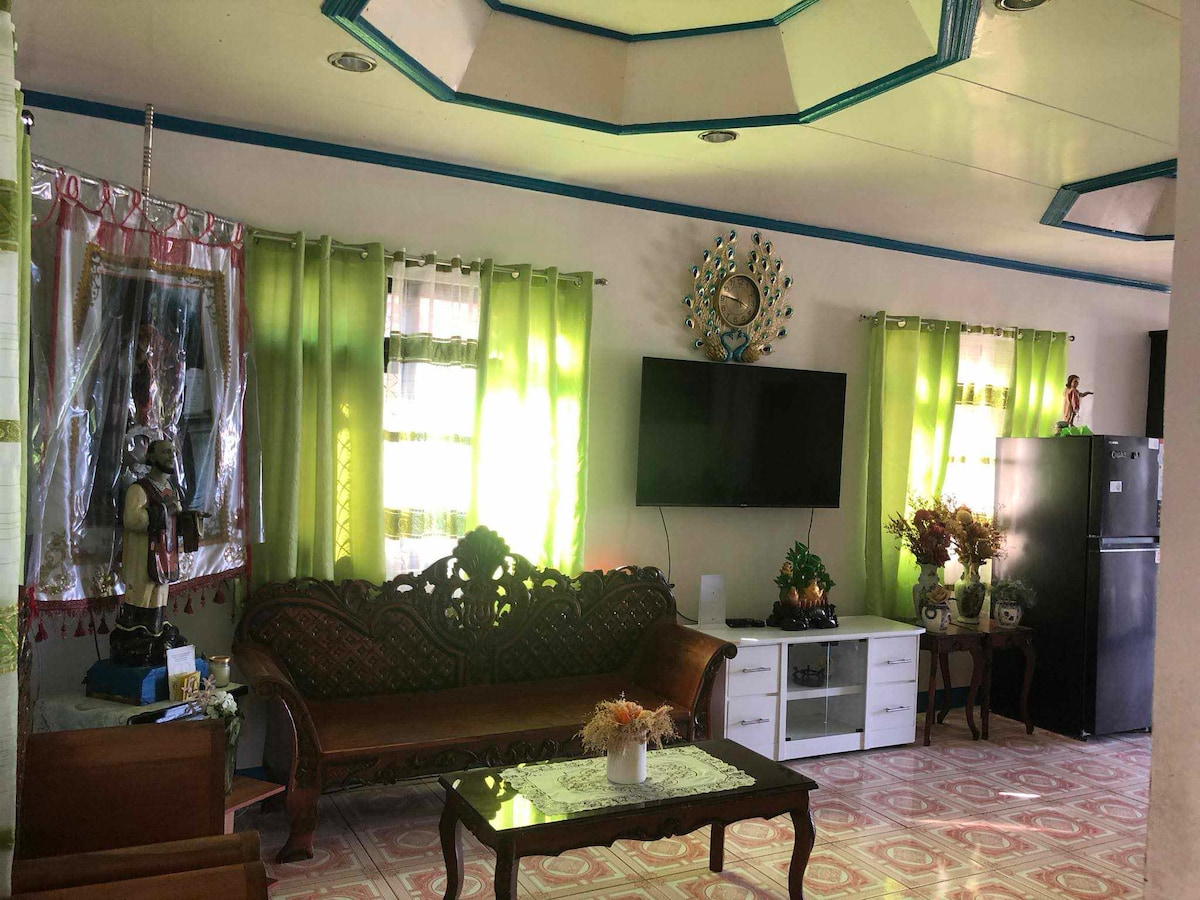 A welcoming living area is highlighted by a well-crafted wooden sofa and a glass-topped coffee table decorated with a small vase of flowers. Light filters through sheer, green curtains, while a television is mounted on a cream-colored wall, accompanied by a sleek refrigerator and a side cabinet.