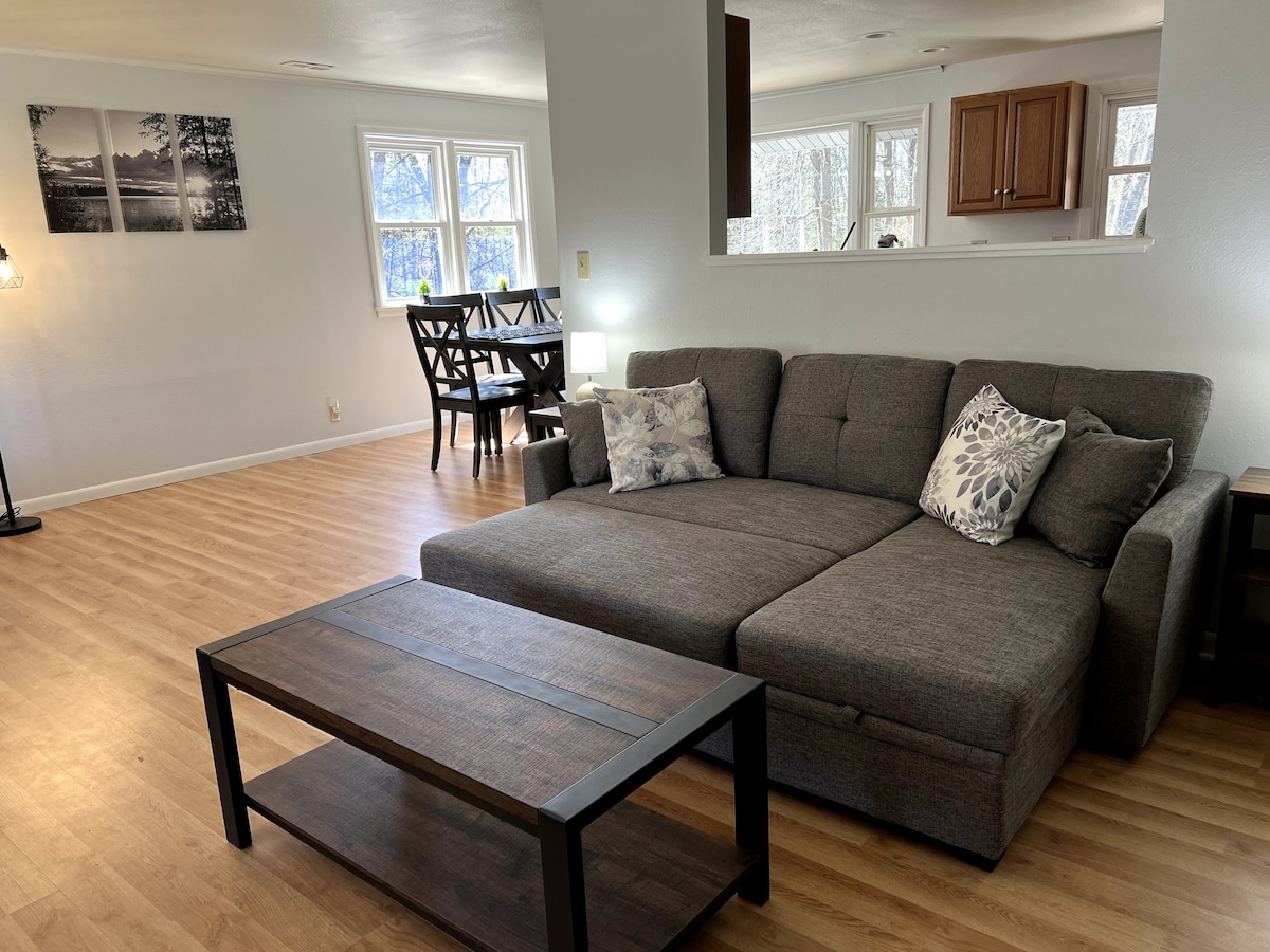 A modern living area features a spacious gray sectional sofa with decorative pillows. A wooden coffee table sits in front, and large windows allow natural light to fill the room. In the background, a dining area with a dark wood table and chairs is visible.
