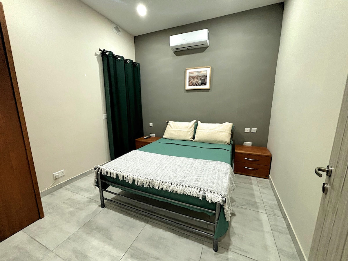 A double bedroom is featured with a spacious bed dressed in green and white linens. The room includes two wooden bedside tables and a framed photograph on the wall. Air conditioning is mounted above, and the room is lit by a ceiling light.