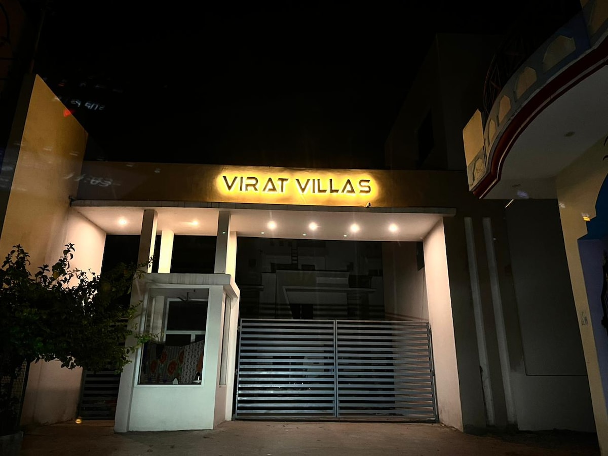 The entrance of Virat Villas is depicted at night, showcasing illuminated signage that reads 'VIRAT VILLAS.' The surrounding area is softly lit, highlighting the modern architecture and a gated entryway with a sleek metal fence.