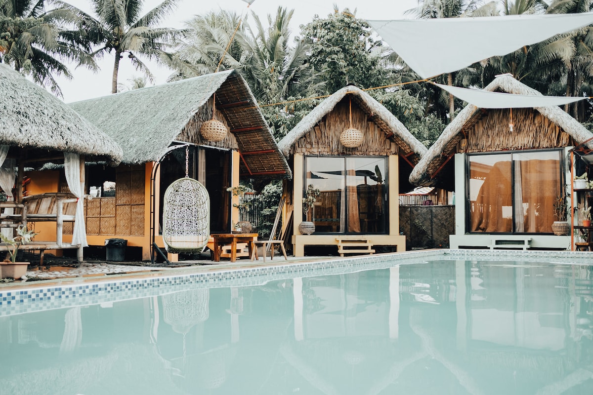 A tranquil pool area is bordered by three cabanas featuring thatched roofs. The still water reflects the surrounding greenery and palms. A hanging chair is positioned near the pool, inviting relaxation in a serene setting.