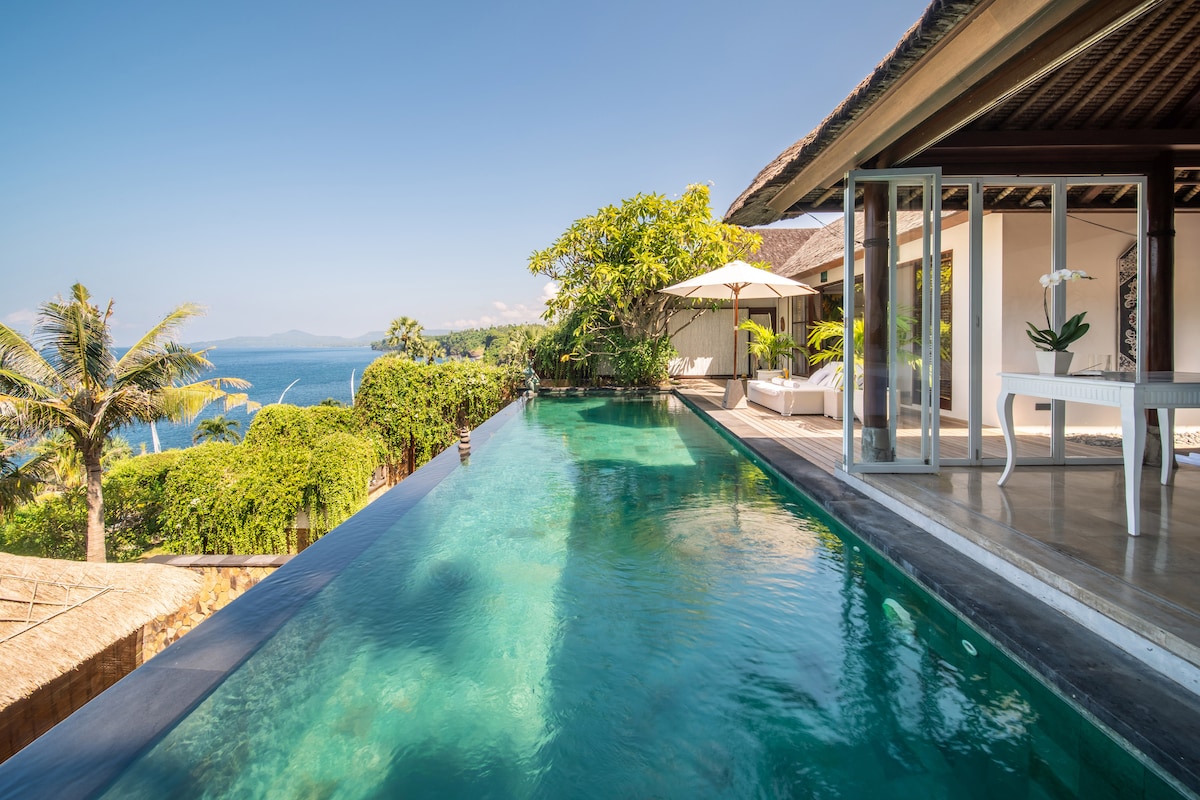 An infinity pool stretches towards the ocean, merging with the serene blue waters. Lush greenery and palm trees provide a natural border, while lounging areas with shaded umbrellas are set nearby, enhancing the tranquil ambiance of the scenic outdoor space.