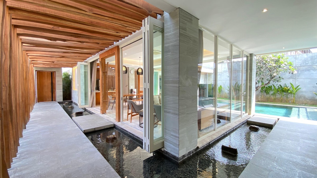 A spacious corridor with natural wood beams leads to an elegantly designed living area. Floor-to-ceiling glass doors allow abundant light and views of the outdoor pool, while the tranquil water feature enhances the serene atmosphere.