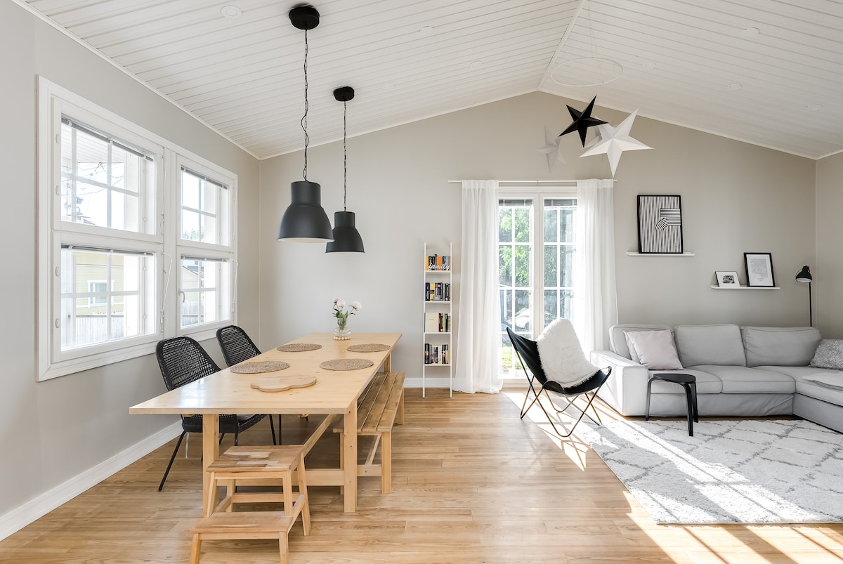 An airy living area features a large wooden dining table with six chairs, complemented by modern pendant lights overhead. A cozy seating arrangement with a gray sofa and decorative pillows is positioned next to large windows, allowing natural light to fill the space.